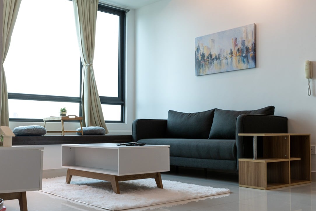 A bright living area is featured with a dark sofa positioned against a large window. A white coffee table rests on a textured rug, while a small shelf stands nearby. Natural light floods the space, highlighting the soft curtains and a piece of wall art.
