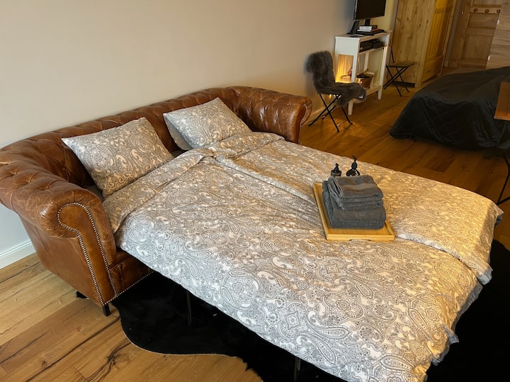 El sofá se extiende en una cómoda cama con sábanas estampadas, que se completa con una bandeja de madera que contiene toallas y artículos de aseo limpios, ubicado en una acogedora sala de estar con suelo de madera.