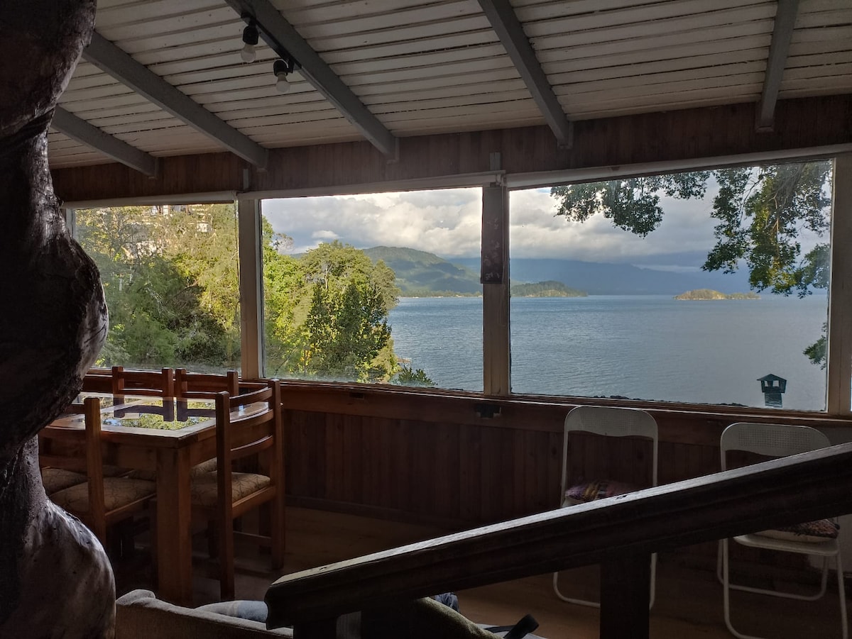 Large windows frame a serene view of the lake and surrounding mountains. A wooden dining table and chairs are positioned in the foreground, creating an inviting spot for meals. Natural light filters through the windows, enhancing the connection to the outdoors.