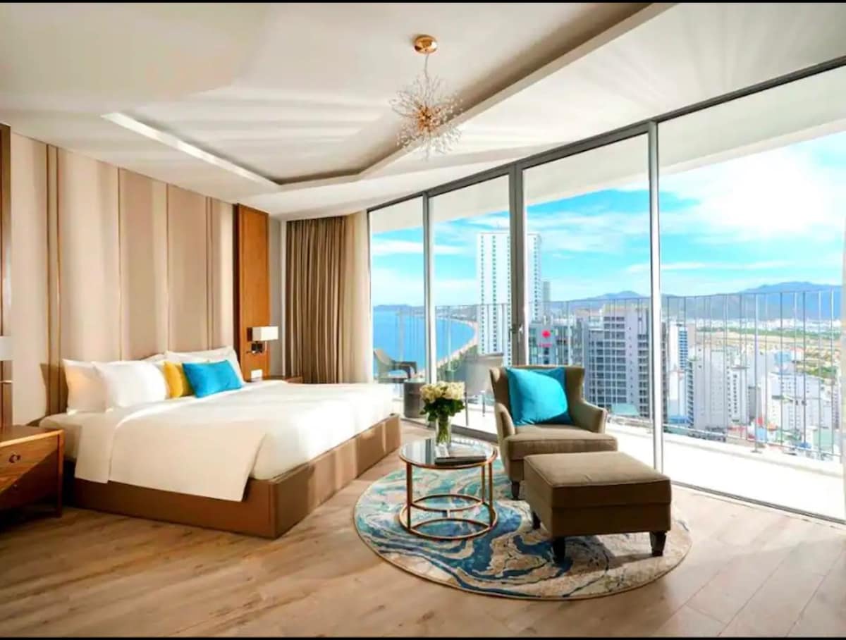 A city view room features a spacious bed with crisp white linens and decorative pillows. A comfortable armchair is positioned next to a round coffee table on a colorful area rug. Large windows flood the room with natural light, showcasing the skyline and ocean beyond.
