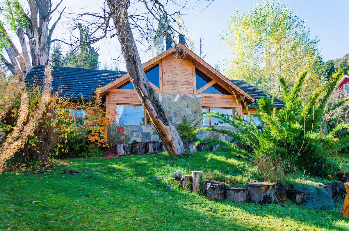 The charming cabin exterior combines native wood and stone, nestled among lush greenery and vibrant trees. A well-maintained lawn envelops the cabin, while large windows provide a glimpse of the inviting interior. Natural elements enhance the tranquil atmosphere of this mountain retreat.