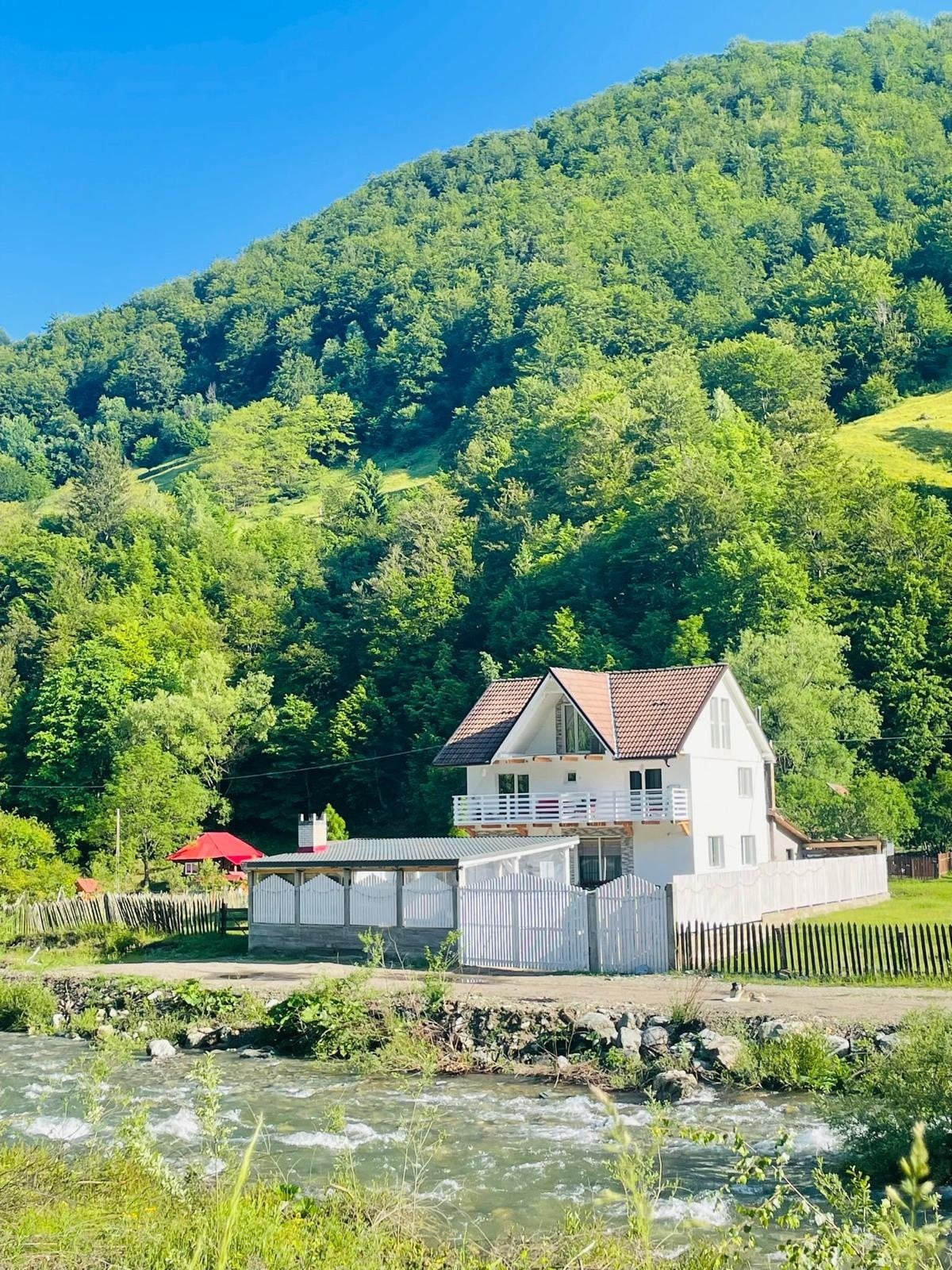 A charming three-story cabin is nestled against a backdrop of lush green mountains. A flowing river runs in front of the property, where a small red gazebo is visible. The exterior of the cabin features a white facade with a sloped roof and multiple balconies.
