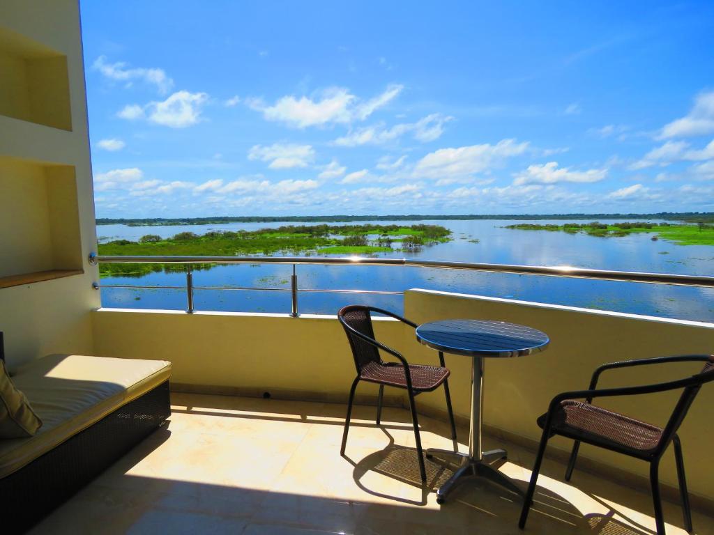 A private balcony offers a circular table and two chairs, overlooking a serene river landscape. Bright blue skies stretch above, reflecting on the water's surface, while lush greenery is visible along the water's edge.