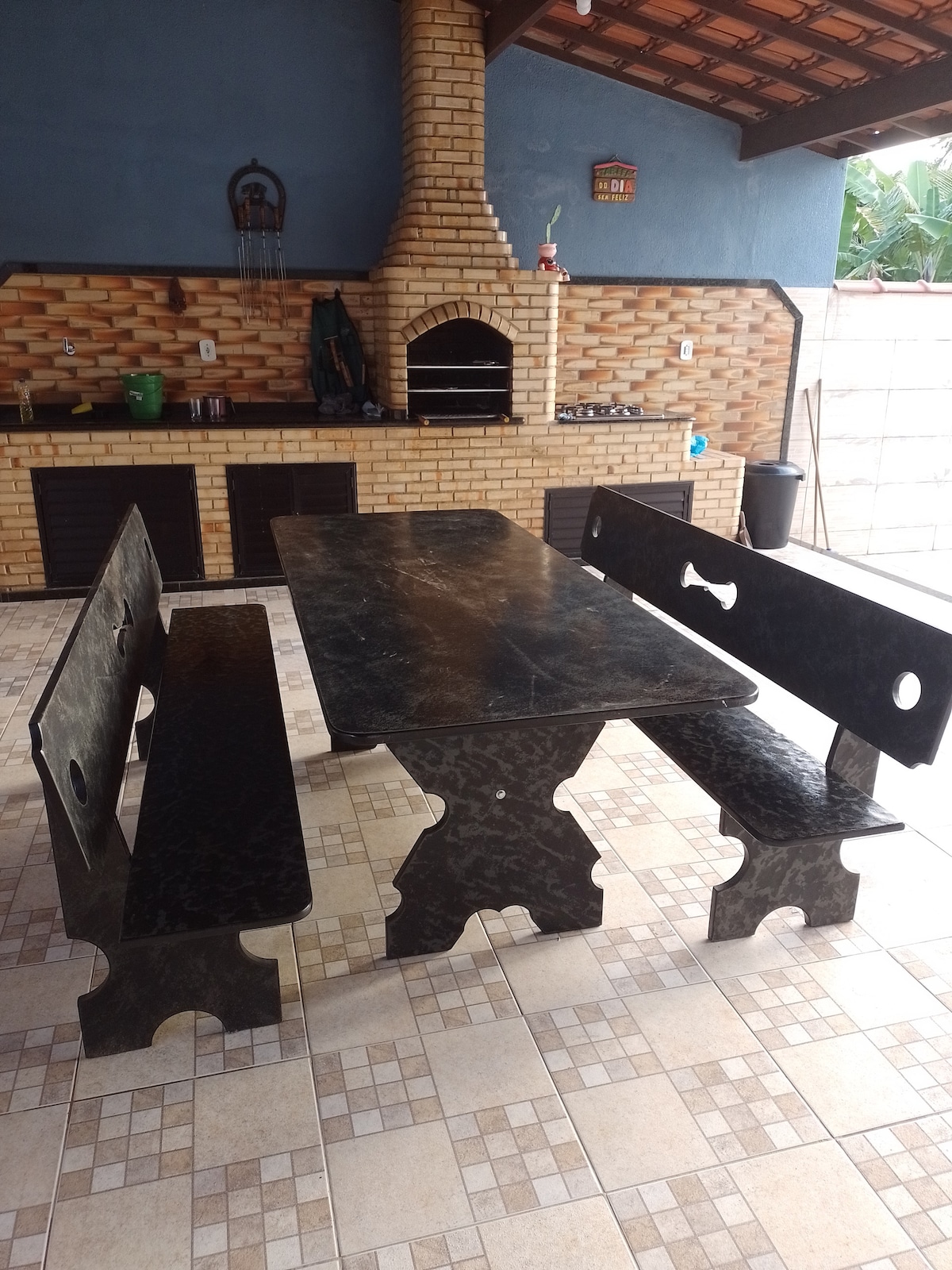 A black stone table with matching benches is positioned in a covered outdoor space. A brick grill is visible in the background, with a rustic brick wall adding texture. The tiled floor features a geometric pattern, enhancing the overall aesthetic of the area.