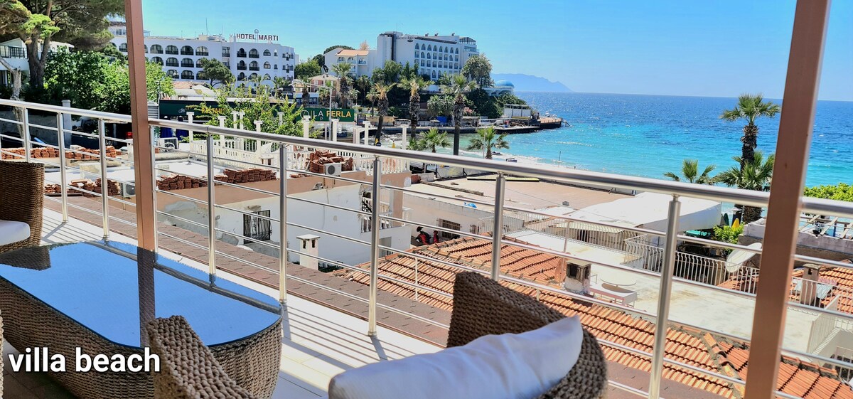 A balcony view showcases a sandy beach and calm sea, with nearby palm trees and coastal buildings. Outdoor seating is arranged around a glass-top table, providing a welcoming space to enjoy the ocean breeze.