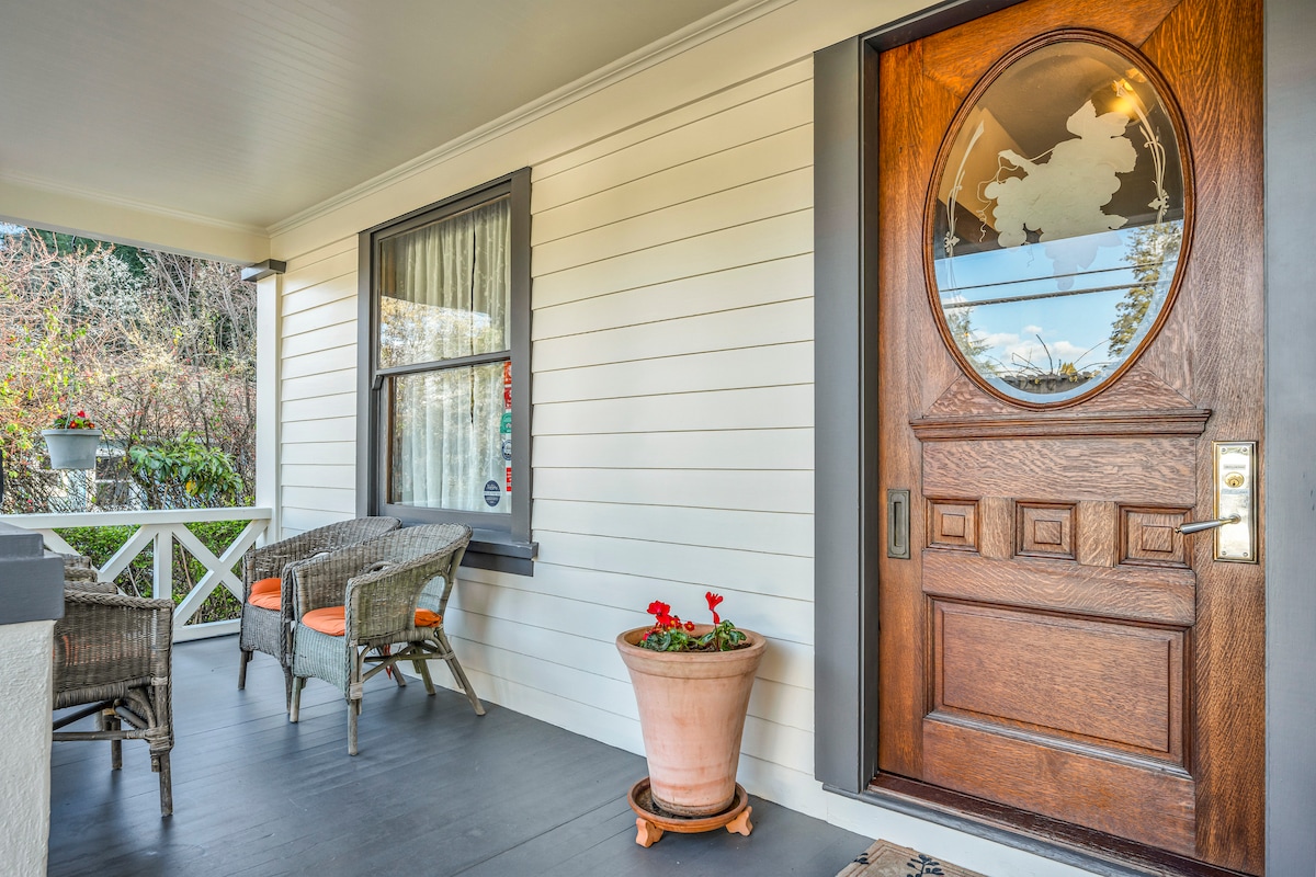 A welcoming front porch features a classic wooden door with an oval glass insert, complemented by a decorative potted plant. Two wicker chairs with orange cushions are arranged around a small table, inviting guests to relax in the peaceful surroundings.
