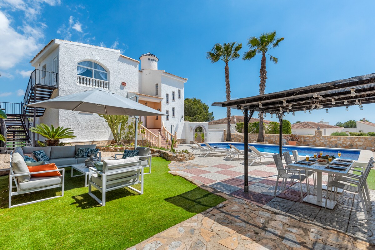 The outdoor area features a well-maintained garden with green grass and stylish lounge seating. A dining setup is positioned under a pergola, while the inviting private pool is visible in the background, surrounded by palm trees and a white villa in the distance.