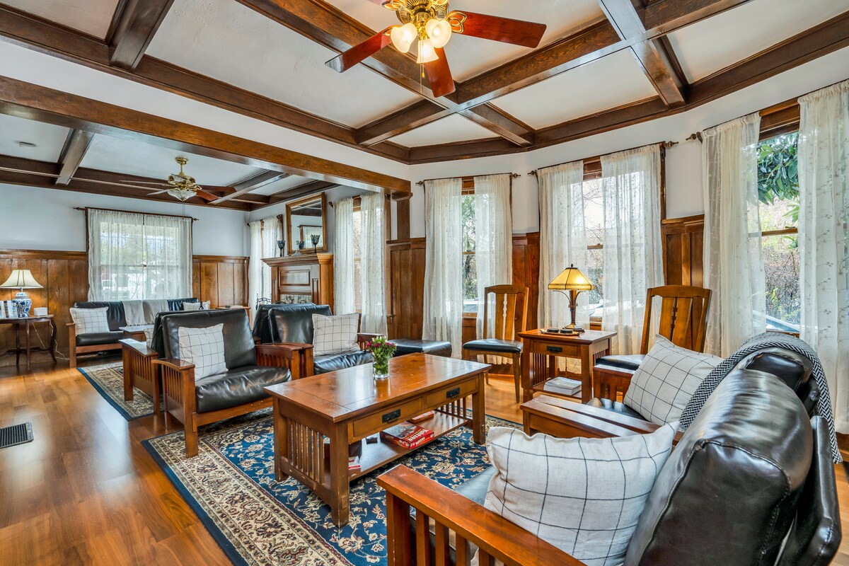 A spacious living area features dark wood beams and light-colored walls, complemented by large windows allowing natural light. Comfortable seating arrangements include leather sofas and plush chairs, surrounding a central wooden coffee table. Decorative elements include a patterned area rug and elegant lighting fixtures.