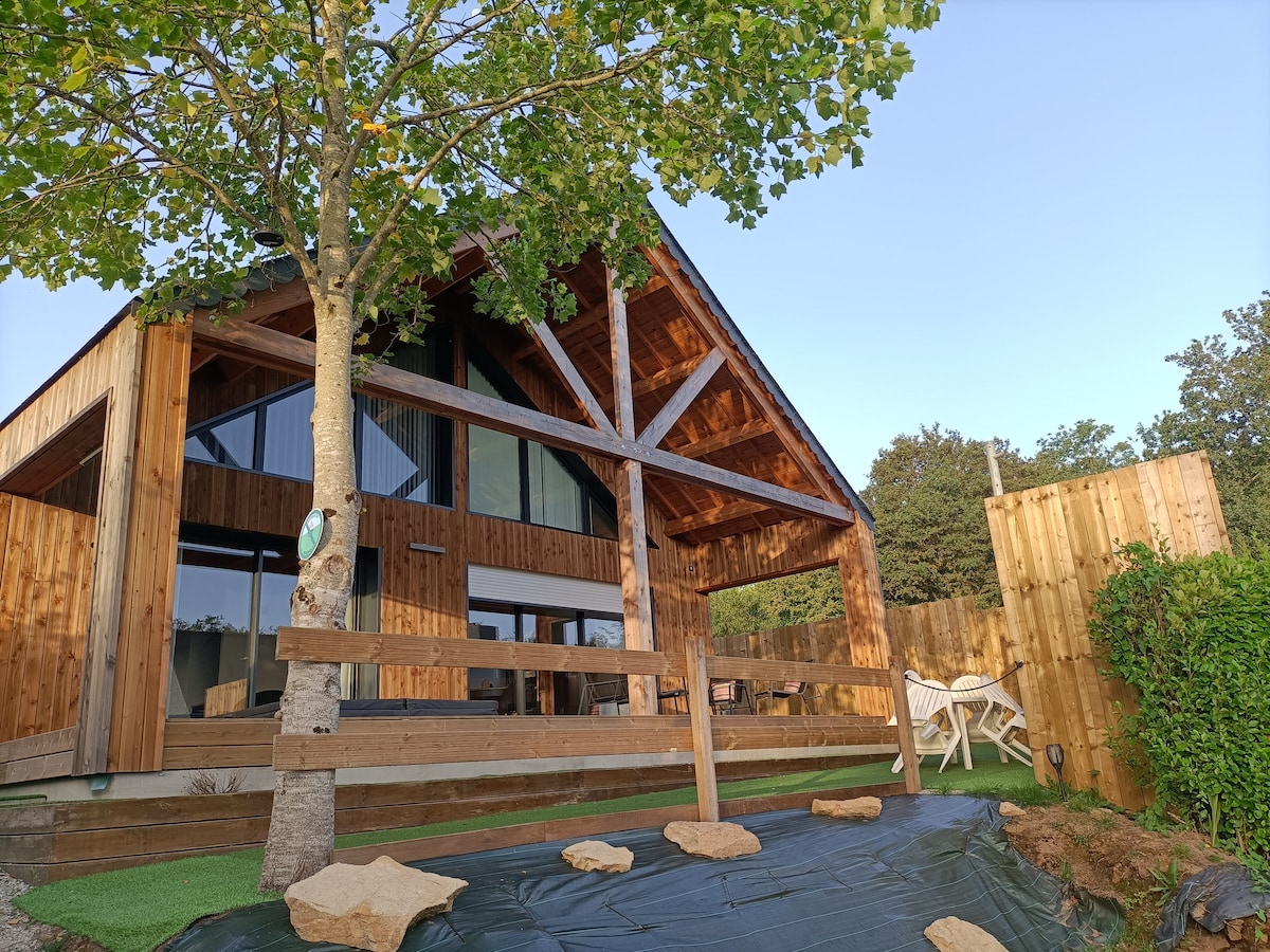 A contemporary chalet with a wooden facade is displayed in the image. The structure features large glass windows and a covered terrace, framed by a prominent tree. Wooden fencing and landscaped greenery create a serene outdoor setting.