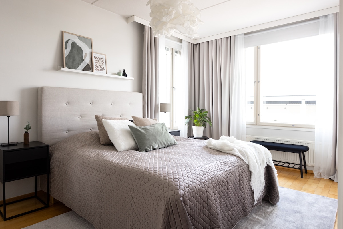 The bedroom features a spacious bed with soft linens adorned in neutral tones. Two lamps are positioned on bedside tables, and a decorative shelf displays framed art. Floor-to-ceiling curtains frame a large window, allowing natural light to brighten the room. A green plant adds a touch of nature.