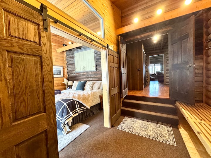 Our primary bedroom is a cozy retreat located down 3 steps from the main floor. A small hallway features a unique wooden slat bench, and across from it is the rustic barn door entrance to the bedroom, creating an inviting and charming atmosphere.
