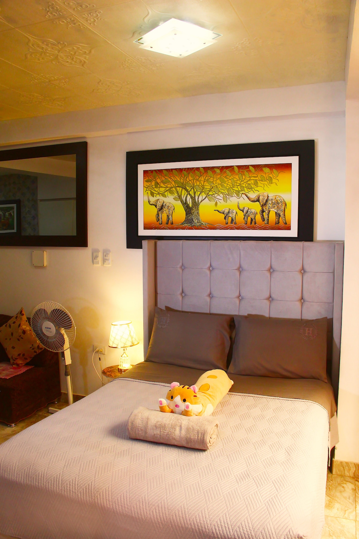 La chambre dispose d'un grand lit avec tête de lit rembourrée et draps gris. Il y a une table de chevet avec une lampe et un ventilateur. Le mur a un tableau avec des éléphants et un arbre. La chambre a un environnement calme et cosy.