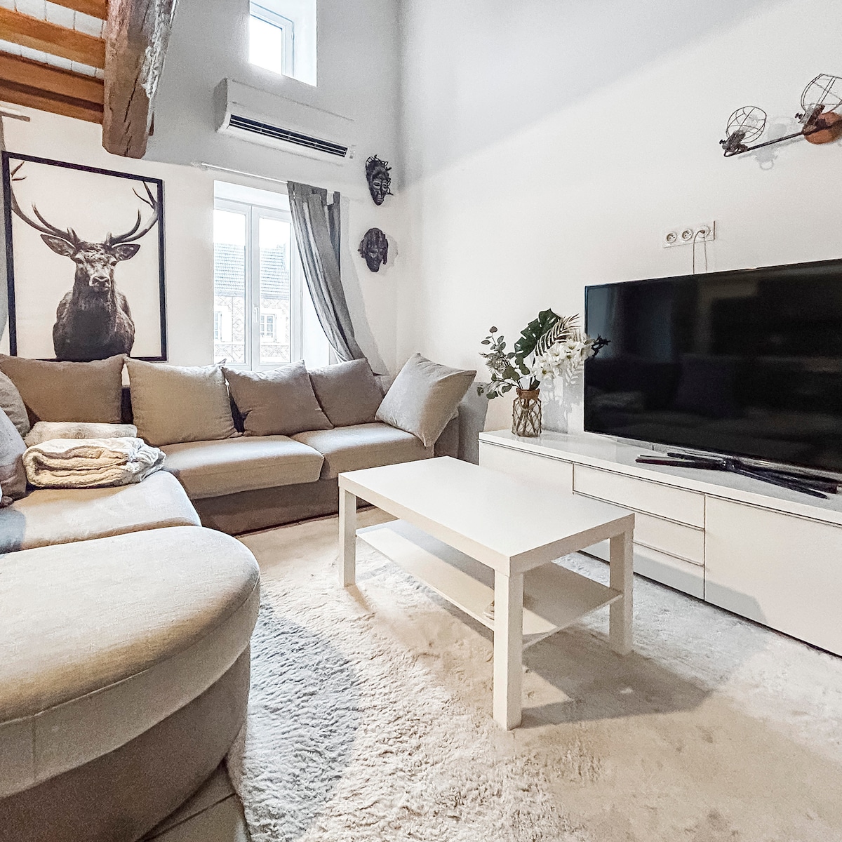 A cozy living room is presented with a large sectional sofa and a round chair, arranged around a white coffee table. A television is positioned opposite the seating, and natural light filters through the windows. Wall decor includes an artwork featuring a deer.
