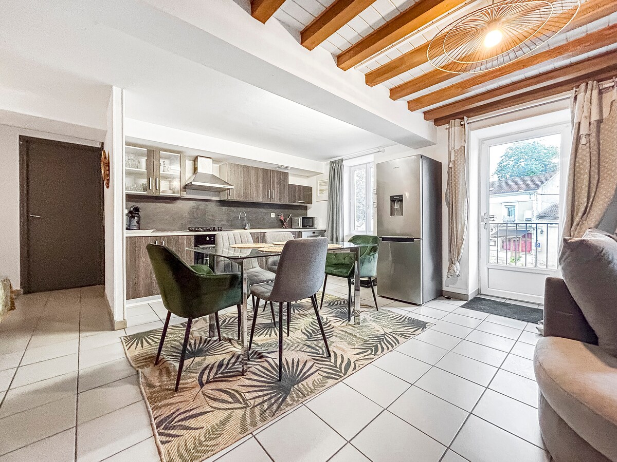 An open kitchen and dining area features a dining table surrounded by four green upholstered chairs. Modern kitchen appliances are visible, with a large refrigerator and sleek cabinetry. Natural light enters through glass doors that lead to a small outdoor space, enhancing the spacious feel.