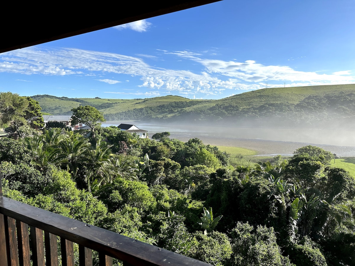 A scenic view of lush green hills and misty landscapes is framed by the wooden deck railing. The area is surrounded by tropical foliage, with patches of open space and a few neighboring houses visible in the distance. Clear blue skies complement the tranquil scene.