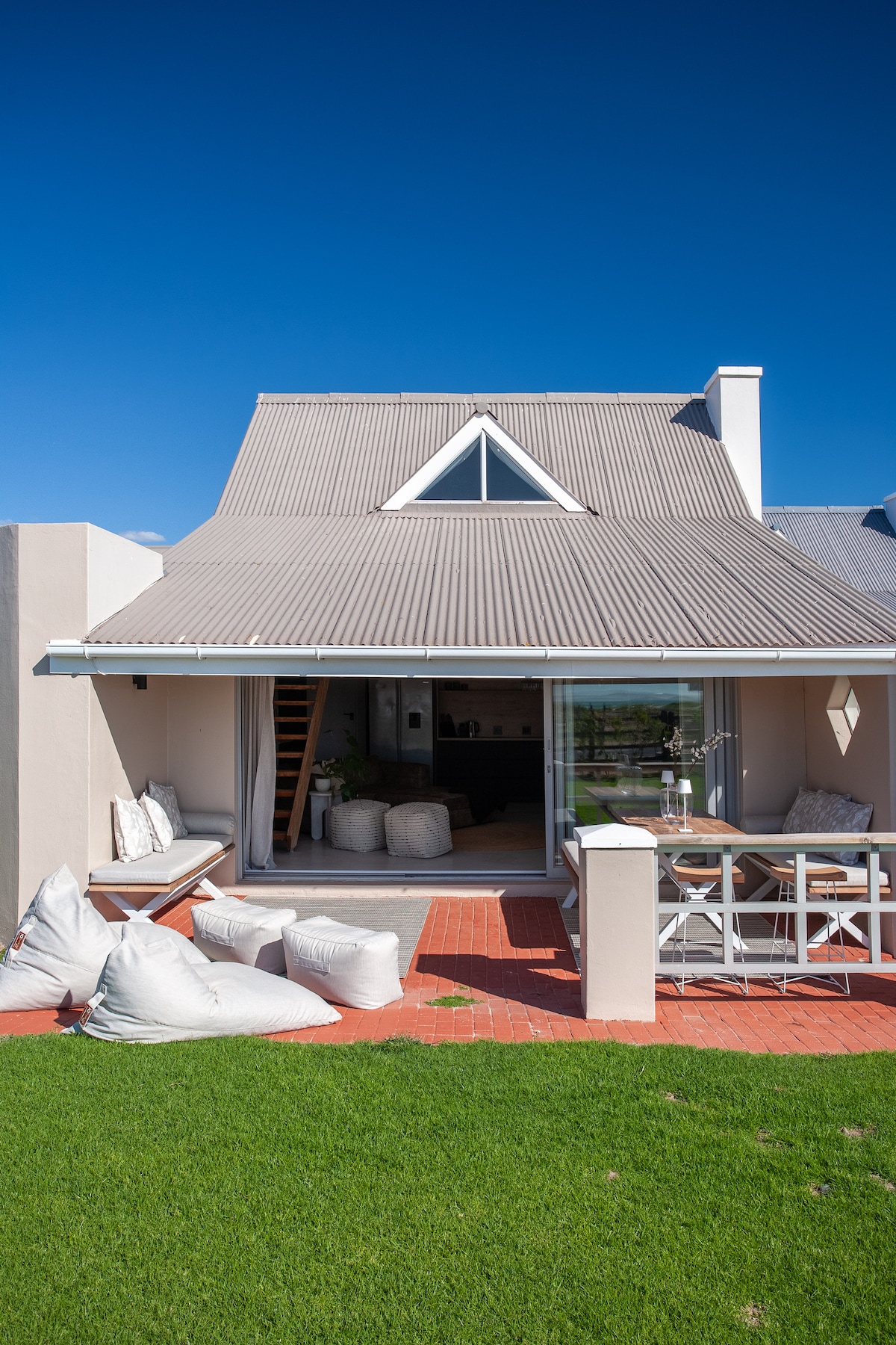 The exterior showcases a charming house with a modern roofing design. The patio features soft seating options, while a wooden table is set for dining. Lush green grass surrounds the area, and large glass doors offer a view into the inviting living space.