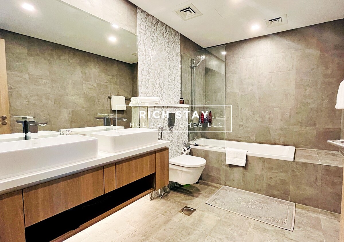 The en-suite bathroom features a spacious layout with dual sinks mounted on a wooden vanity. A glass-enclosed walk-in shower and a separate bathtub are included, with modern fixtures complementing the sleek tile design. Fresh towels and toiletries are neatly arranged for guest convenience.
