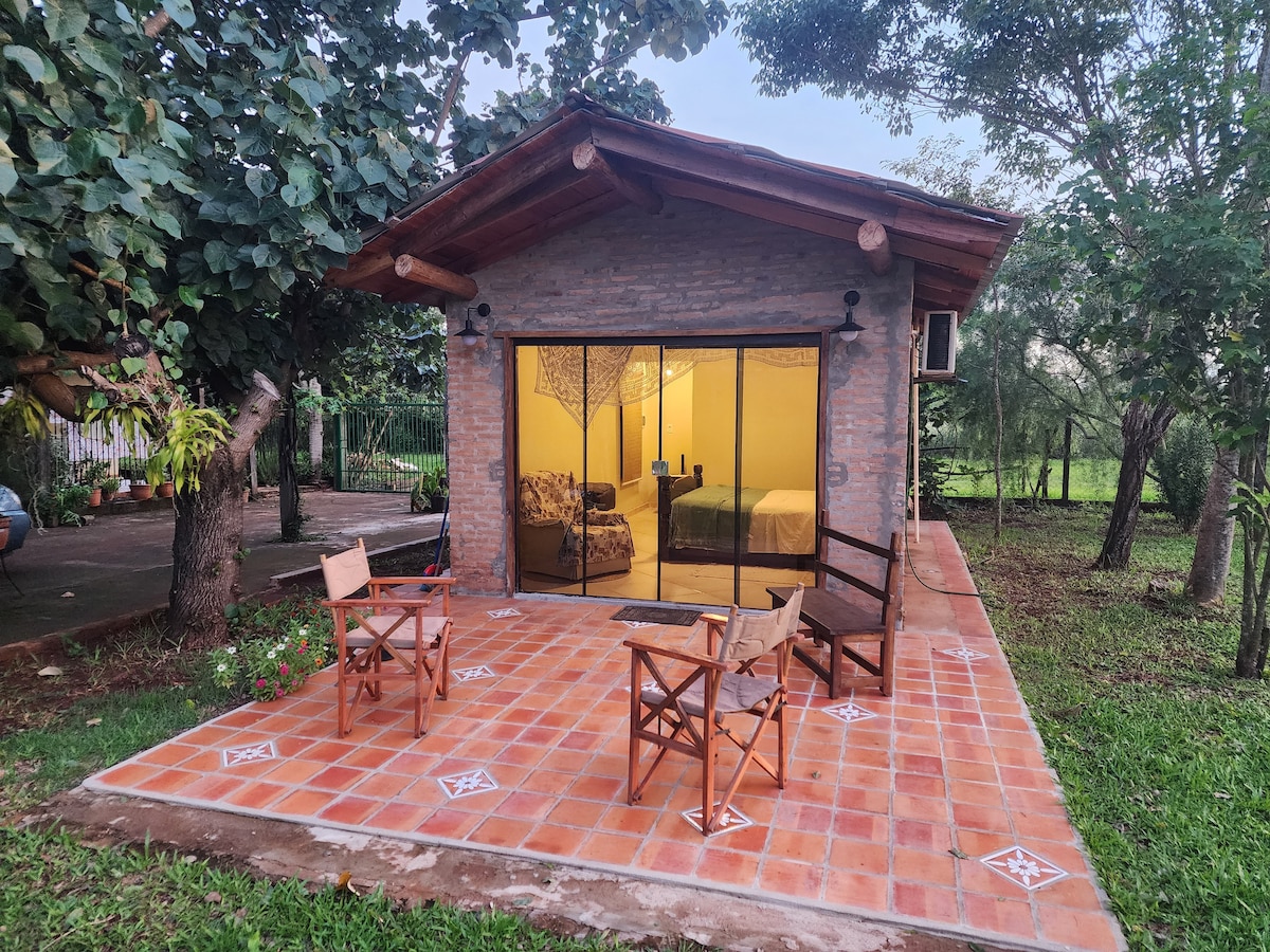 An outdoor patio is shown, featuring a tiled floor and four wooden chairs arranged around a small table. Large glass doors provide a view into the interior, where a cozy living space is visible with comfortable seating and a spacious bed. Lush greenery borders the area.