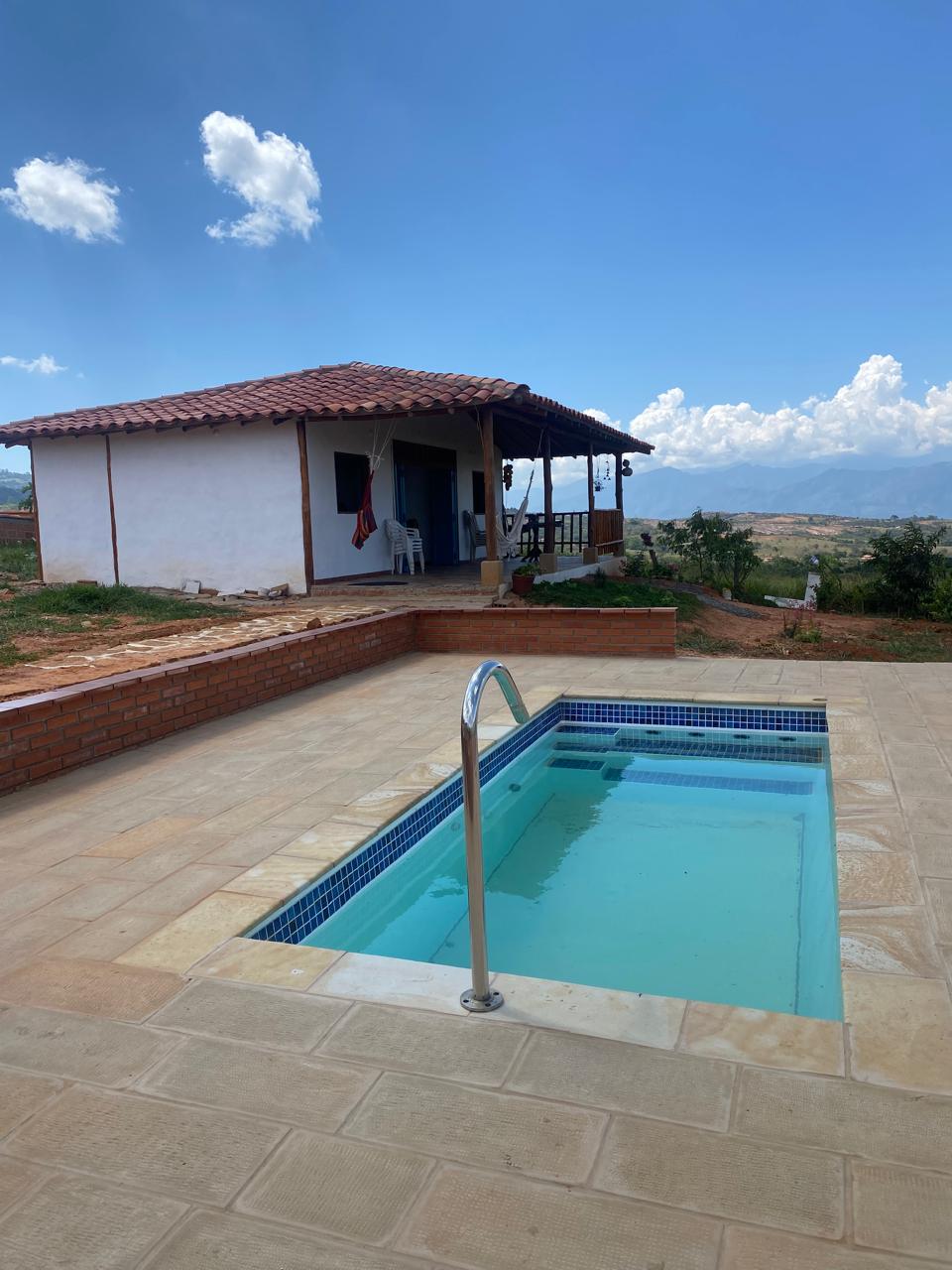 A small house is situated near a tranquil swimming pool, surrounded by scenic landscapes. The pool, featuring a steel railing, is set on a tiled patio that extends toward the house, which has a traditional roof and open porch accessible for relaxation.