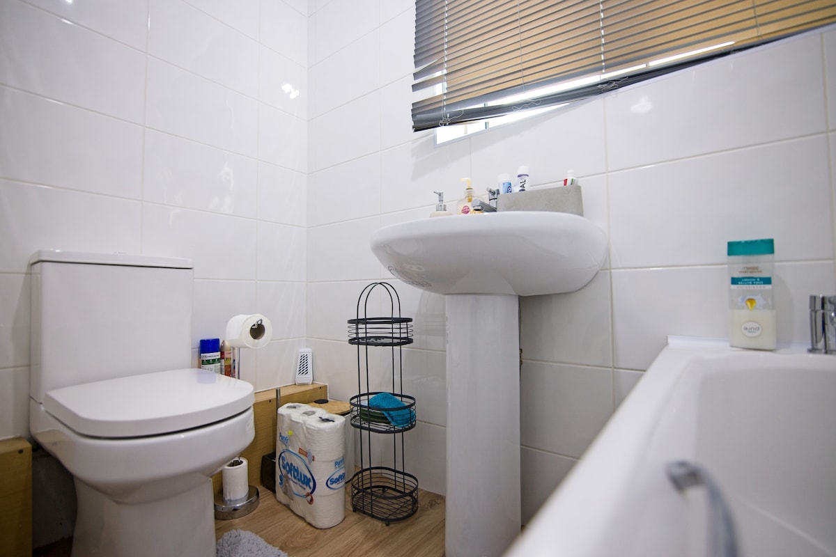 The bathroom features white tile walls and a clean aesthetic. A compact toilet is positioned next to a modern pedestal sink. Above the sink, natural light enters through a partially covered window, while toiletries and essentials are organized on nearby shelves.
