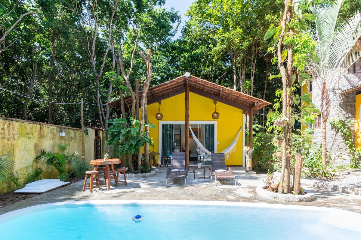 A vibrant yellow chalet is framed by lush greenery, featuring a covered porch with two lounge chairs and a hammock. A spacious pool reflects the surrounding trees, and a wooden table with chairs is positioned nearby, creating a relaxing outdoor retreat.