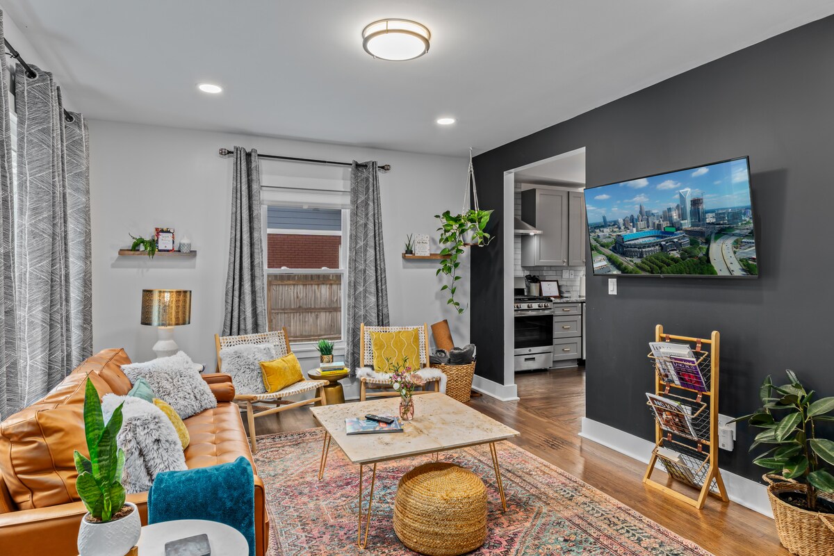 A modern living area features a stylish leather sofa and cozy seating options, with a coffee table at the center. A large wall-mounted TV displays an urban view, while decorative plants and artwork add character. Soft lighting enhances the inviting atmosphere.