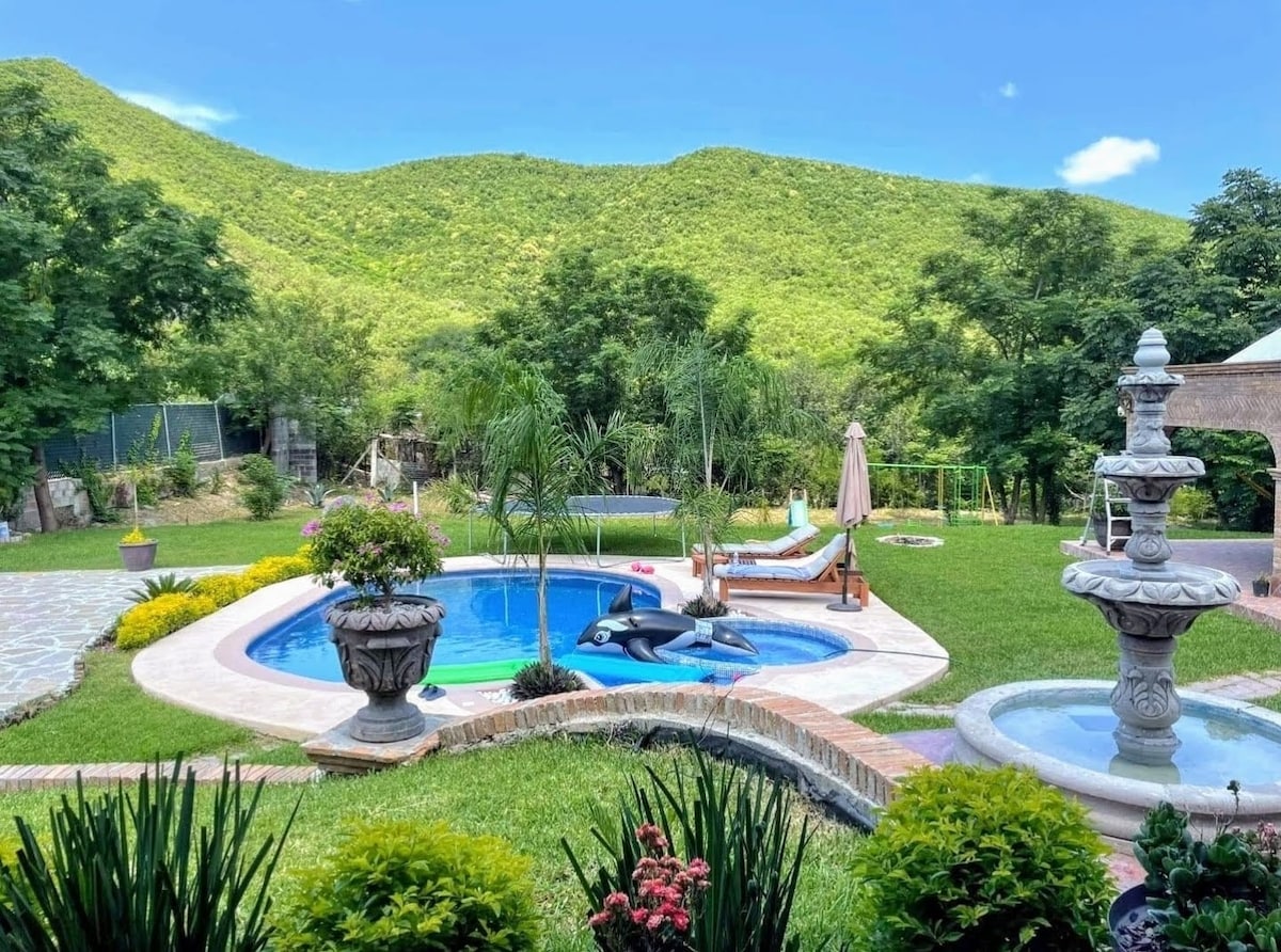 The outdoor area features a swimming pool surrounded by lush greenery, with mountains visible in the background. Relaxing loungers and a shaded area are positioned near the pool. Vibrant flowers and decorative fountains enhance the tranquil garden setting.
