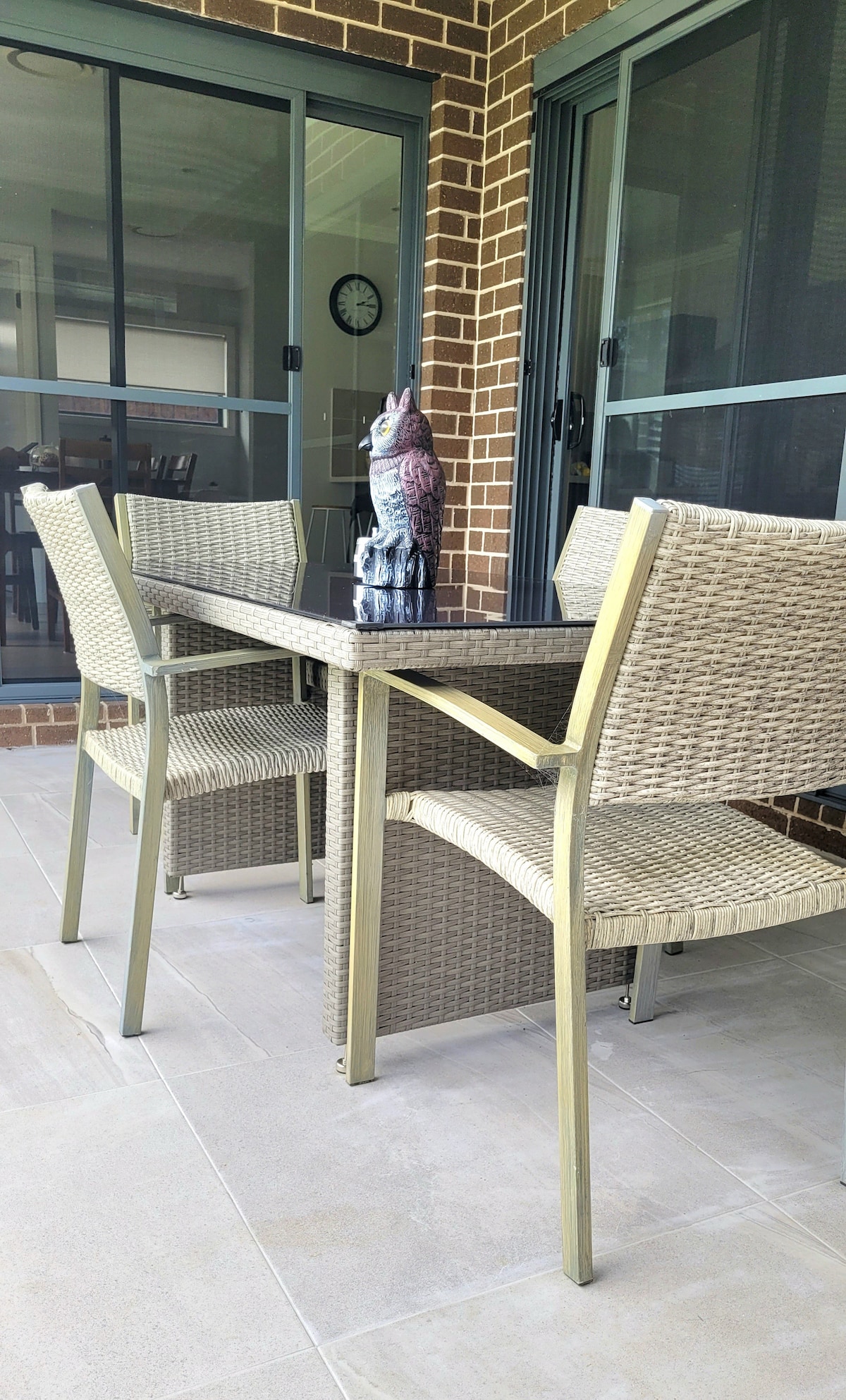 An alfresco dining area is depicted, featuring a glass-top table surrounded by four woven chairs. A decorative owl statue is centered on the table, with sliding glass doors in the background providing access to the indoor living space.