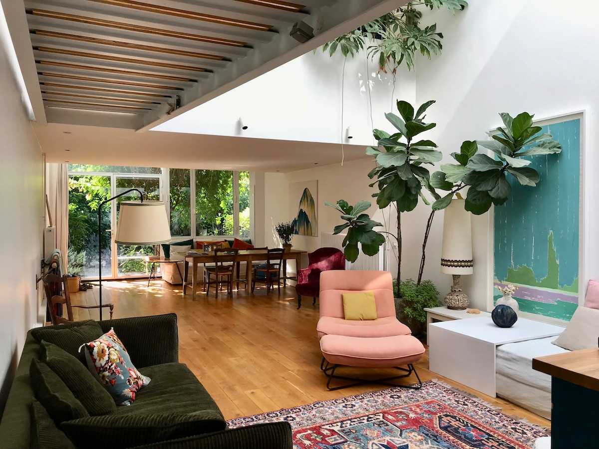 A spacious living area features a cozy green sofa and a pink lounge chair, surrounded by vibrant indoor plants. The polished wooden floor complements the light streaming in through large glass windows that overlook a green garden. A dining table is visible in the background.