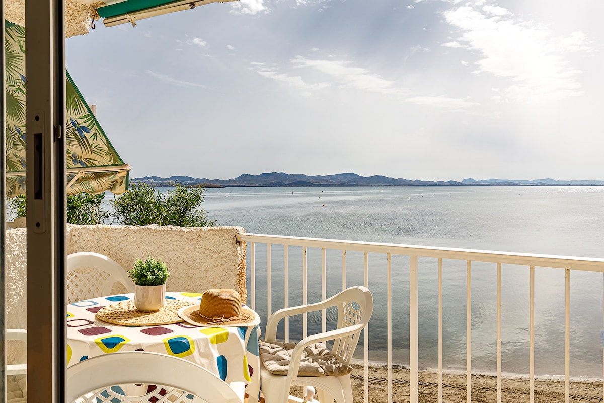 A beachfront terrace is visible, featuring a round table with a colorful tablecloth and two white chairs. The calm water of Mar Menor reflects the soft light, while distant hills create a serene backdrop. An umbrella provides shade, enhancing the relaxing space.