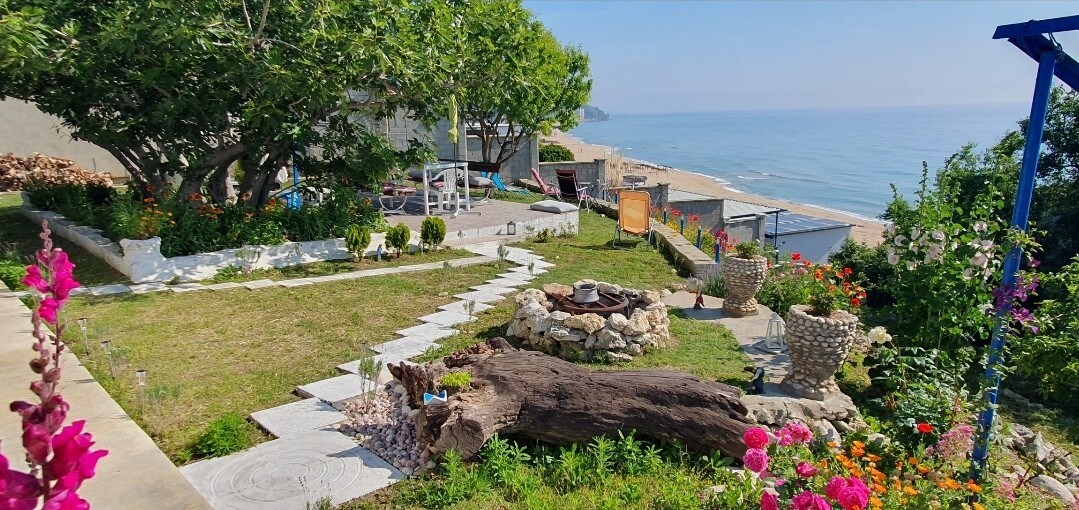 A spacious garden is adorned with vibrant flowers and greenery, leading towards a coastal view. Seating areas, a fire pit, and lounge beds are positioned throughout the space, offering spots to relax while overlooking the sea. The beach is visible in the background.