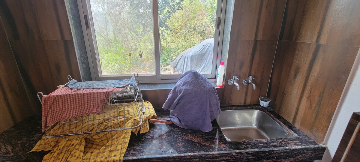 The kitchen area features a granite countertop with a stainless steel sink. An organized drying rack holds dishcloths, while a covered item rests nearby. Natural light enters through a large window, revealing a view of the greenery outside.