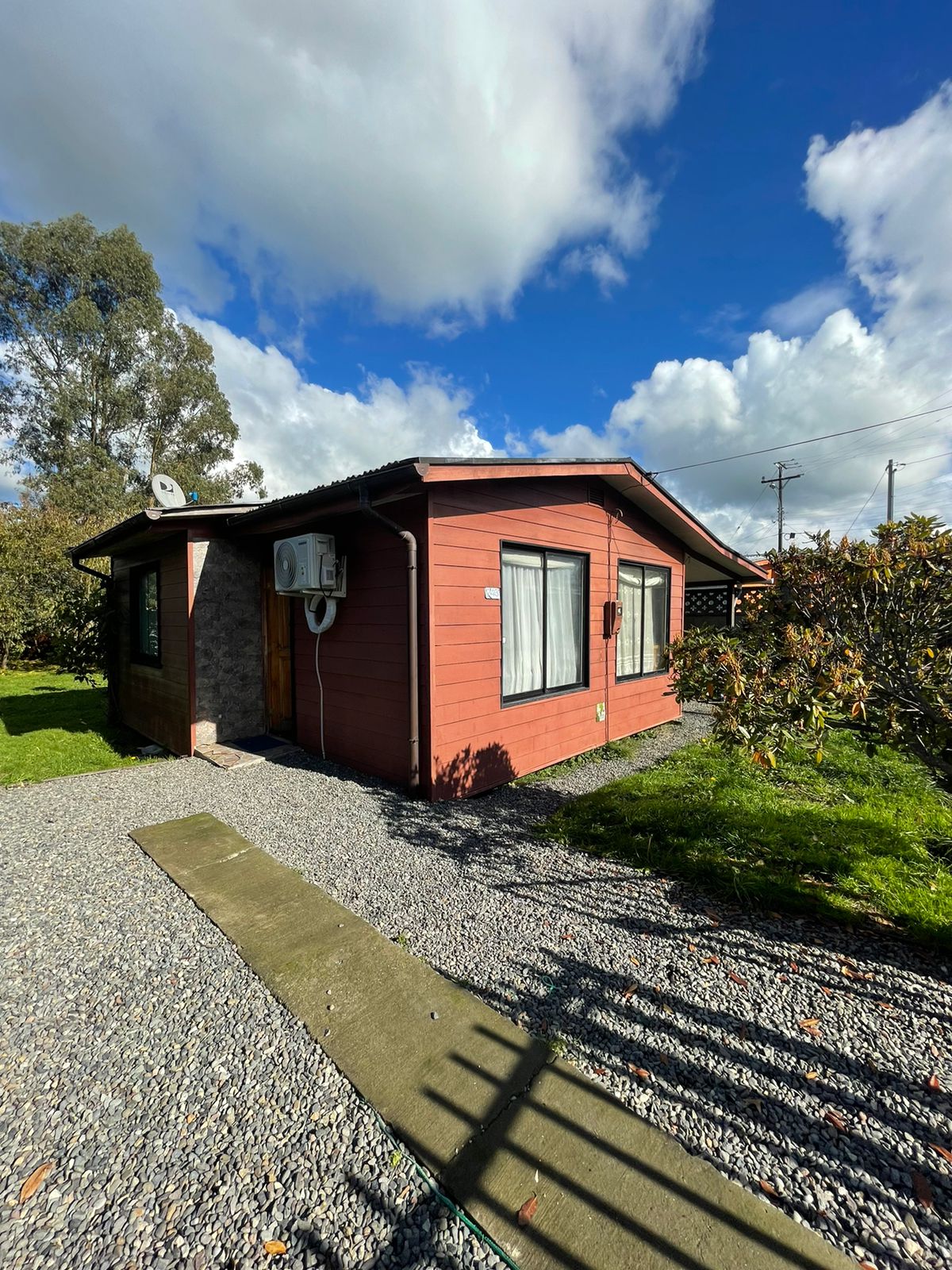 The exterior of the cabin is visible, featuring a red wooden facade and large windows. A gravel pathway leads to the entrance, surrounded by green grass and shrubs. The sky is partly cloudy, enhancing the natural setting.