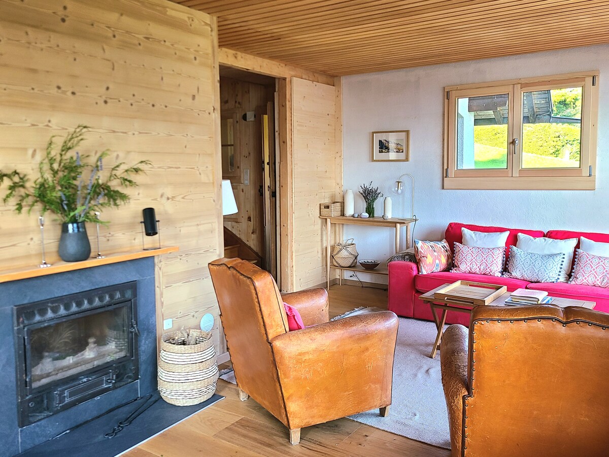A cozy living room features a fireplace with a dark stone surround and wooden accents. Two leather armchairs are positioned beside a small table, and a red sofa adorned with decorative pillows adds a pop of color. Natural light fills the space through a modest window.