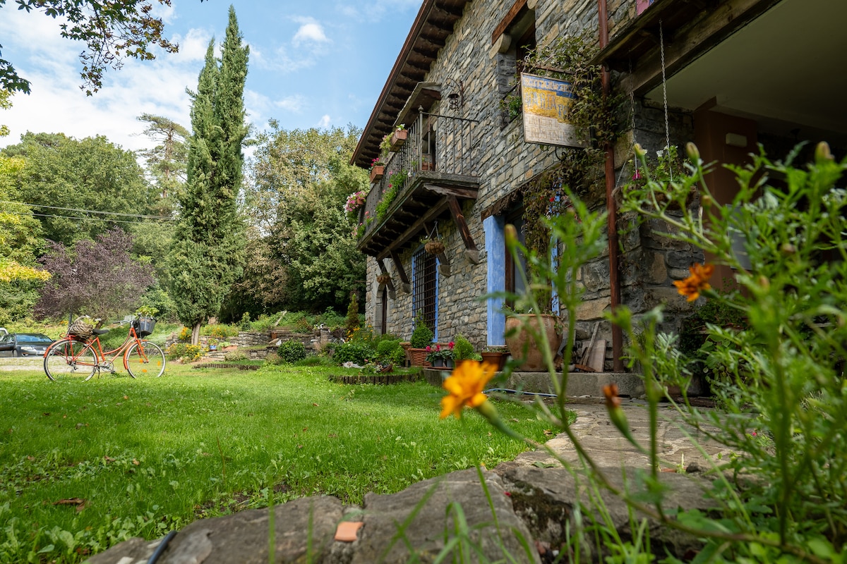 The exterior of the stone building is framed by a lush green lawn and vibrant flowers. Two bicycles are positioned nearby, suggesting opportunities for outdoor exploration. A variety of trees and plants create a natural backdrop, enhancing the tranquil setting.