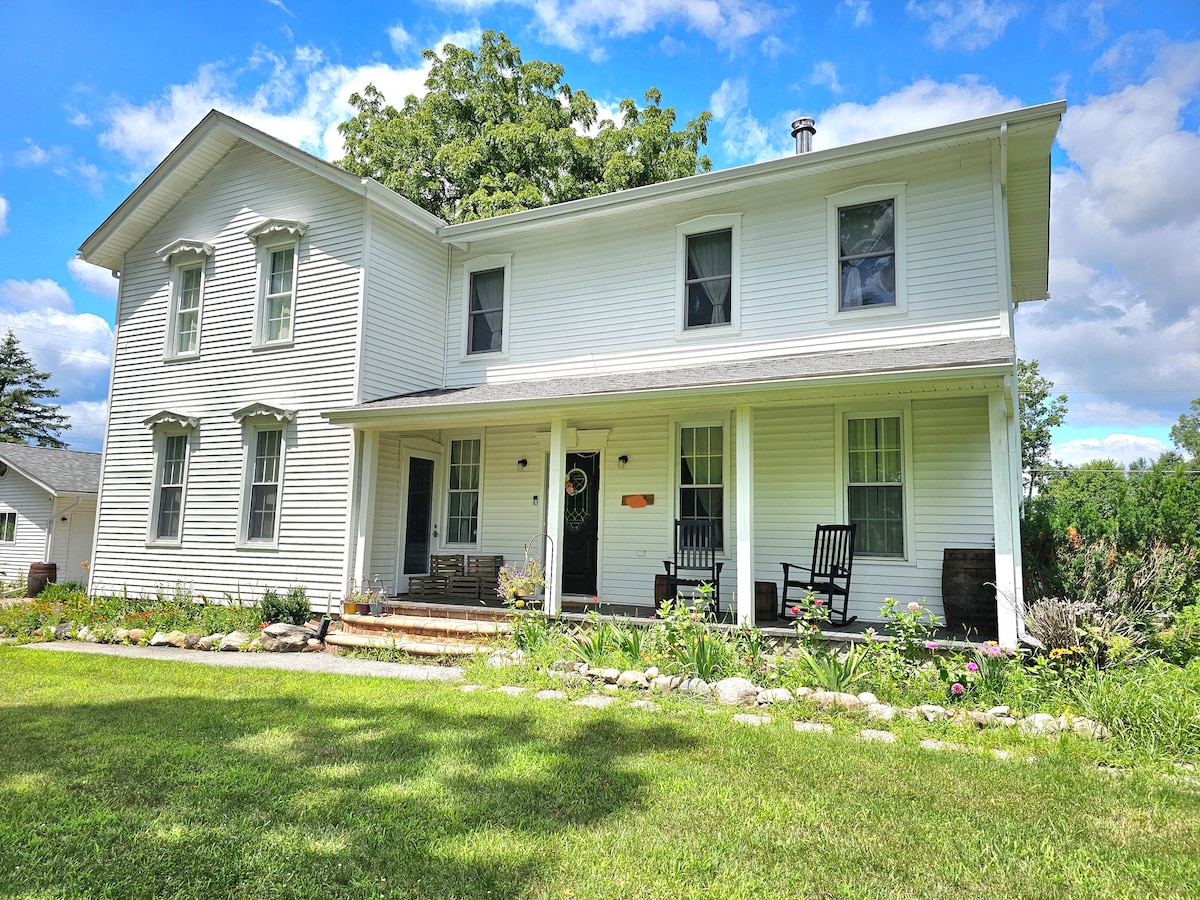 The charming farmhouse features a neat exterior with white siding and a welcoming front porch. Rocking chairs are positioned for relaxation, while a landscaped garden with flowering plants enhances the front yard. Large windows allow natural light to fill the interior spaces.