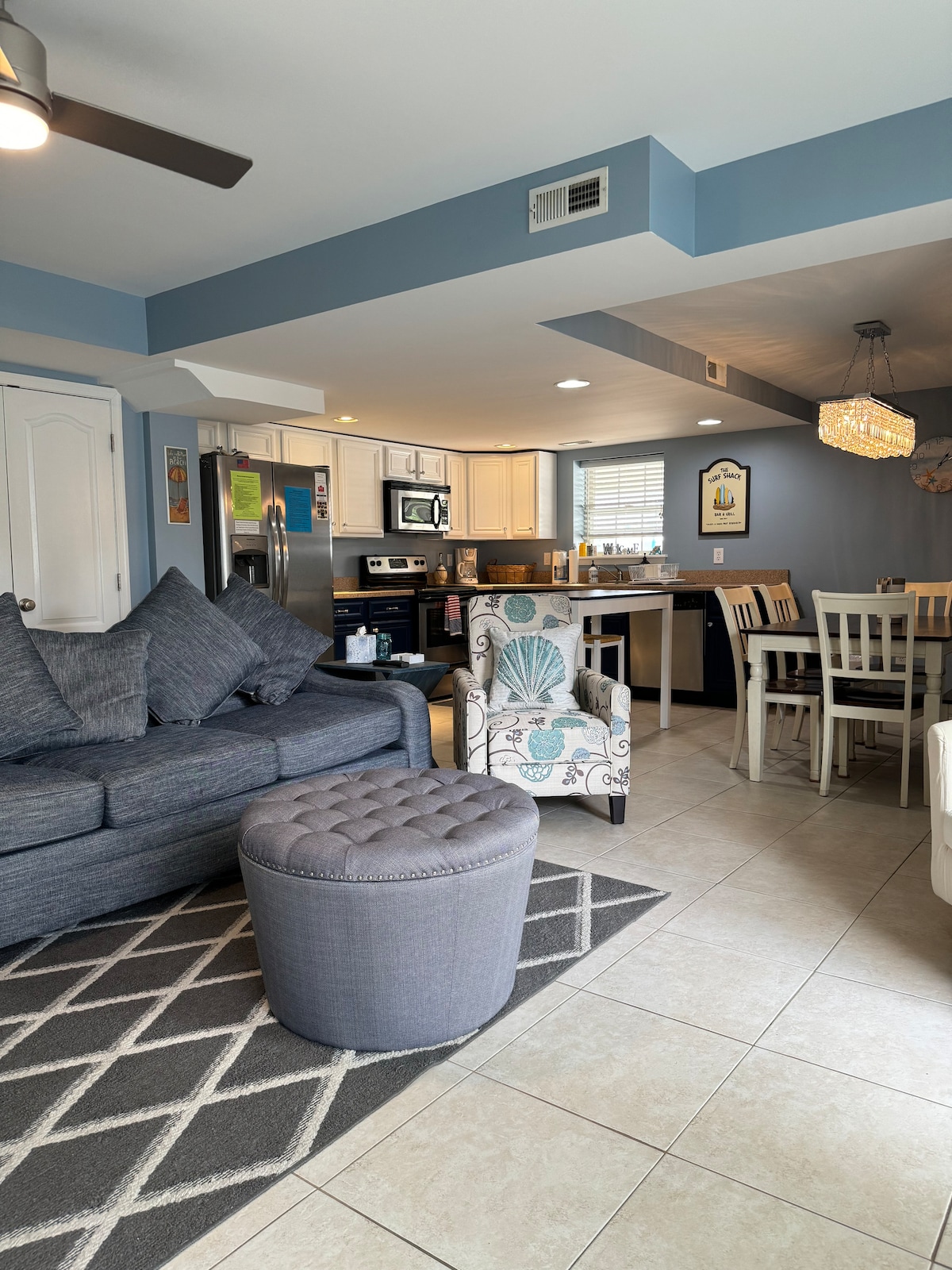 A modern living area features a plush sofa adorned with decorative pillows. A compact ottoman sits nearby on a textured rug. The open kitchen showcases stainless steel appliances and a breakfast bar, while a dining table accommodates chairs for shared meals. Natural light filters through the windows.