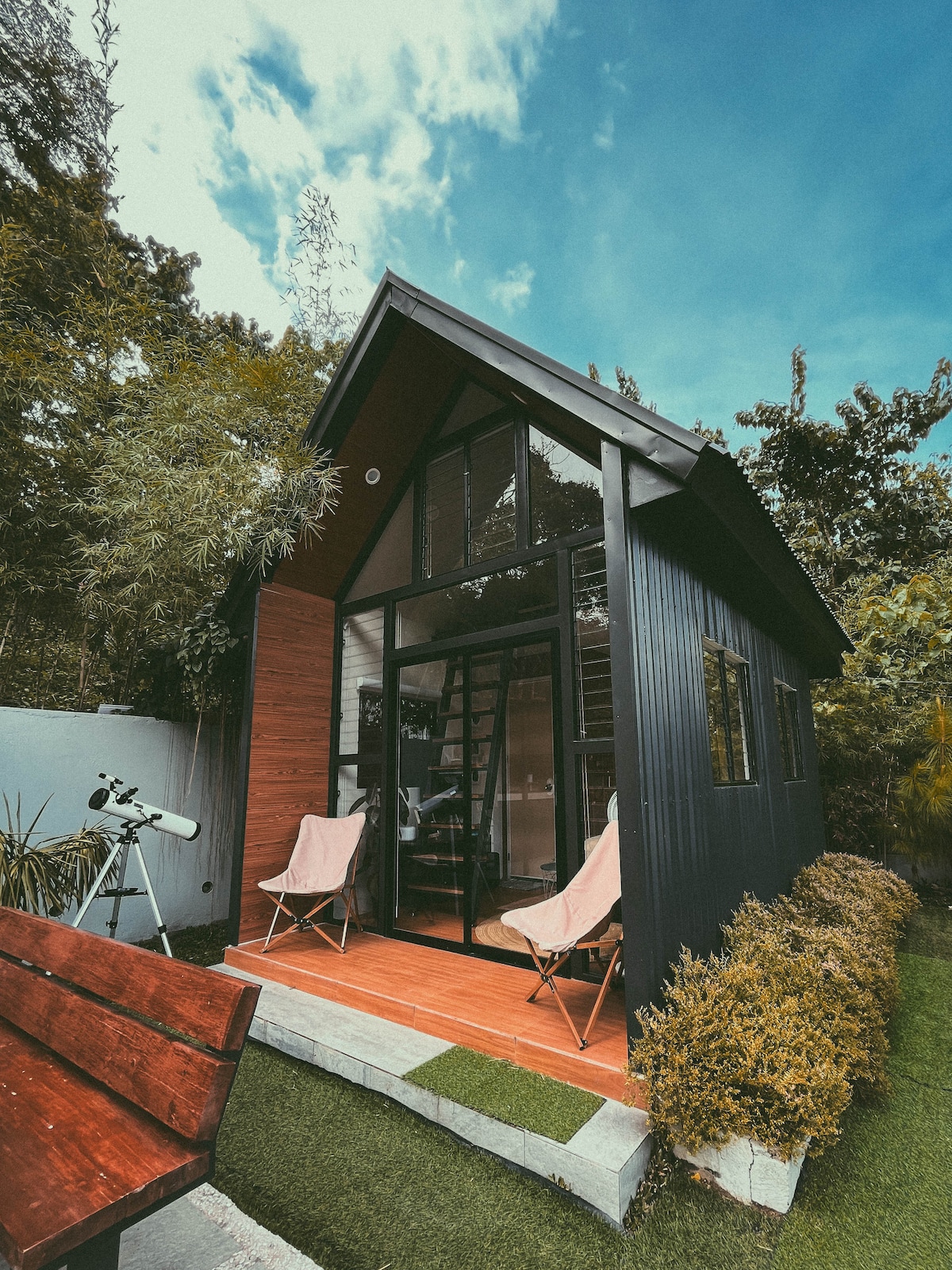 The cabin exterior presents a modern design with sharp angles, featuring dark black panels and warm wooden accents. Two lightweight chairs are positioned outside, inviting relaxation. Lush greenery surrounds the space, complemented by a telescope resting nearby for stargazing opportunities.
