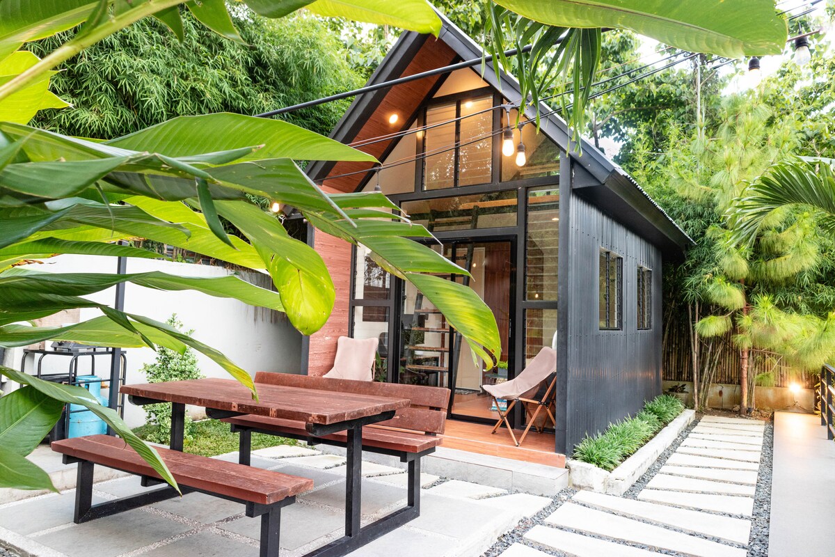 An inviting outdoor area features a rustic picnic table with benches surrounded by vibrant greenery. The black cabin, with large windows, is visible in the background, showcasing a blend of modern design and natural elements. Soft lighting accents the pathway leading to the entrance.