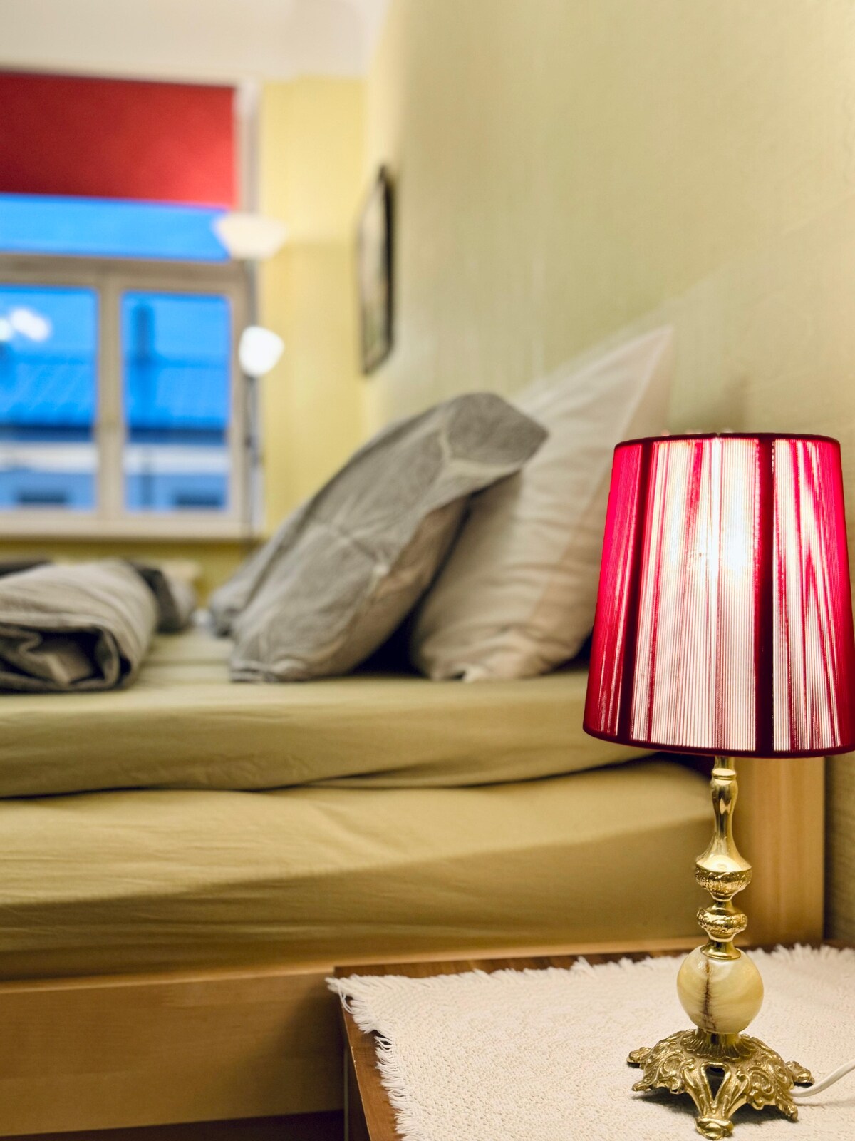 A cozy sleeping area features a spacious bed with light-colored bedding and multiple soft pillows. A small bedside table holds a red-striped lamp, providing a warm glow. The room is illuminated by natural light streaming through a window with red curtains.