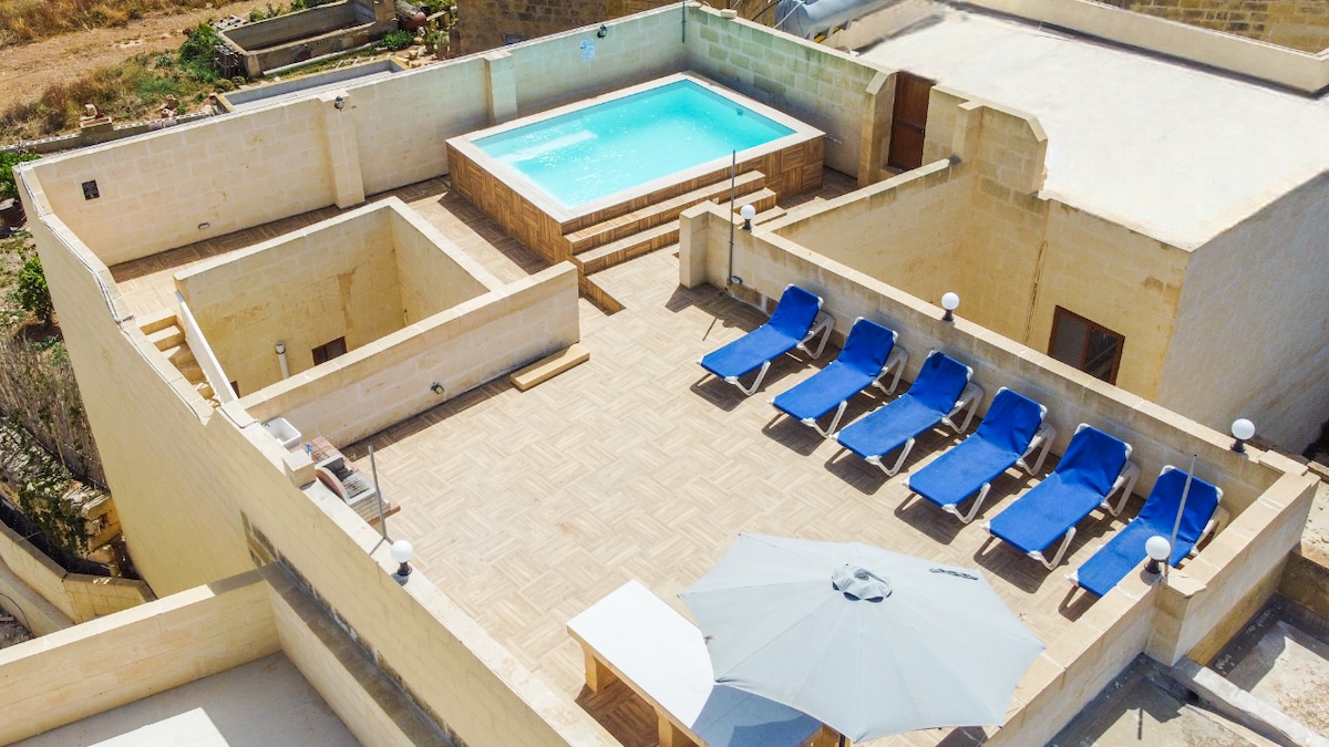 A rooftop terrace is presented with a private swimming pool flanked by sun loungers and a shaded area featuring an umbrella. The surrounding deck is constructed of light-colored stone, offering ample space for relaxation. A lower terrace area is also visible.