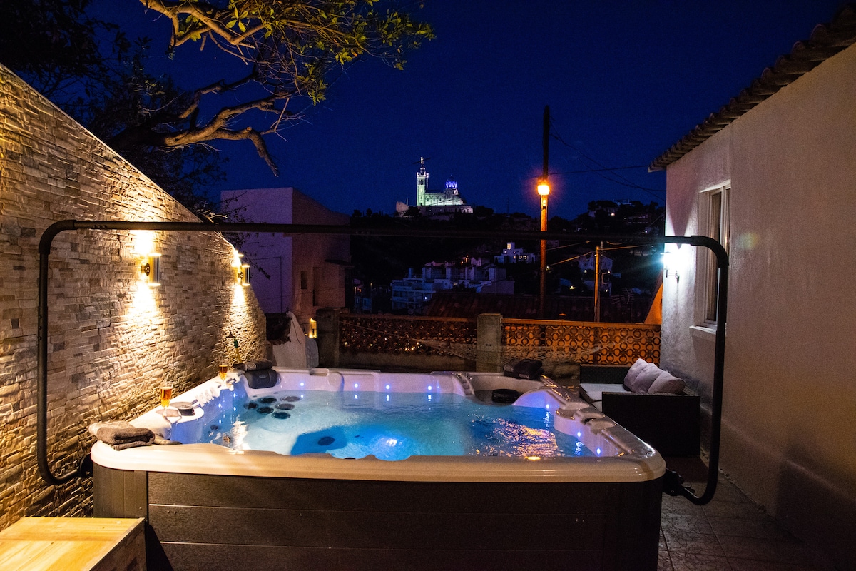 A spacious jacuzzi is illuminated against a stone wall, with soft lighting creating a relaxing ambiance. In the background, the silhouette of Notre-Dame de la Garde is visible, adding a scenic touch to the outdoor space at night.