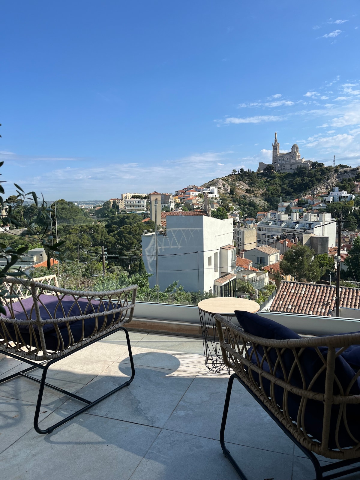 A view from a terrace featuring two stylish chairs, overlooking a panoramic landscape. The Basilica of Notre-Dame de la Garde is prominently positioned on a hill in the distance, surrounded by residential buildings and greenery under a clear blue sky.