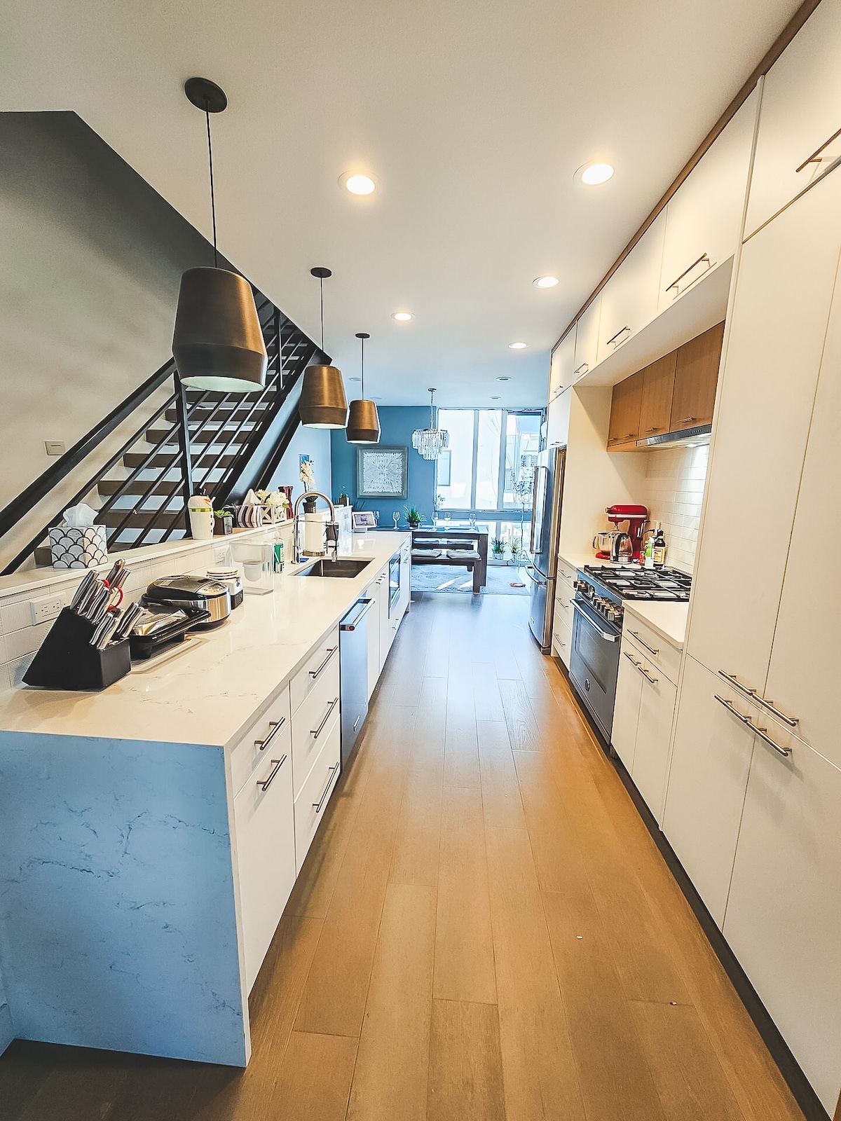 A modern kitchen features white cabinetry and a marble countertop, with a chef's grade gas stove and stylish appliances visible. Pendant lights cast a warm glow over the space. A doorway opens into a light-filled living area, enhancing the kitchen’s inviting and functional layout.