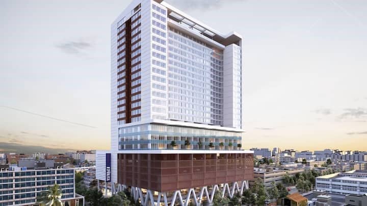 A modern high-rise building is portrayed against a clear sky, showcasing a sleek façade with a combination of glass and metal. The structure is supported by a distinctive base elevated on stilts, highlighting its contemporary architectural design and urban setting.