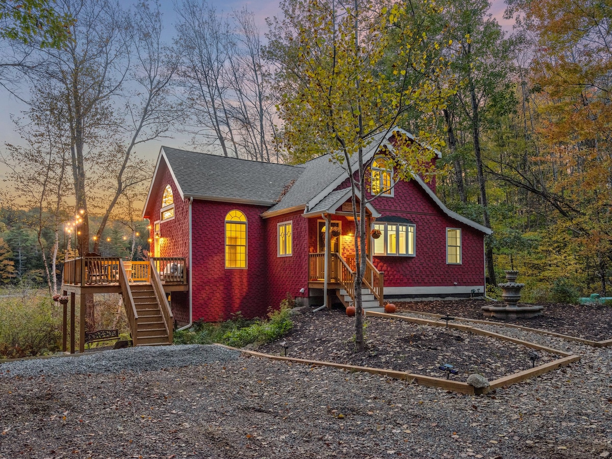 A charming red cottage is framed by vibrant autumn foliage, with warm lights glowing through its windows. The inviting front porch is accessed by a wooden staircase, leading to a serene outdoor setting surrounded by nature.