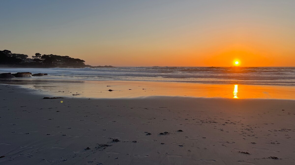A tranquil beach scene captures the sun setting on the horizon, casting warm orange and golden hues across the sky. The calm waves gently lap against the shoreline, reflecting the vibrant colors on the wet sand, creating an inviting atmosphere.