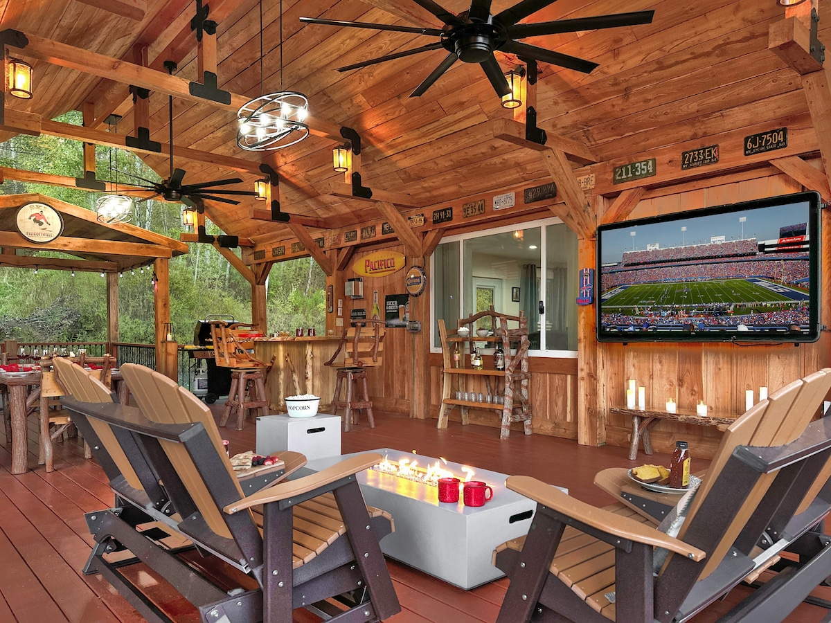 A spacious covered deck features timber framing, with comfortable seating arranged around a gas fire table. A large television is mounted on an adjacent wall, and warm lighting highlights the wooden structure. The atmosphere invites relaxation and social interaction.