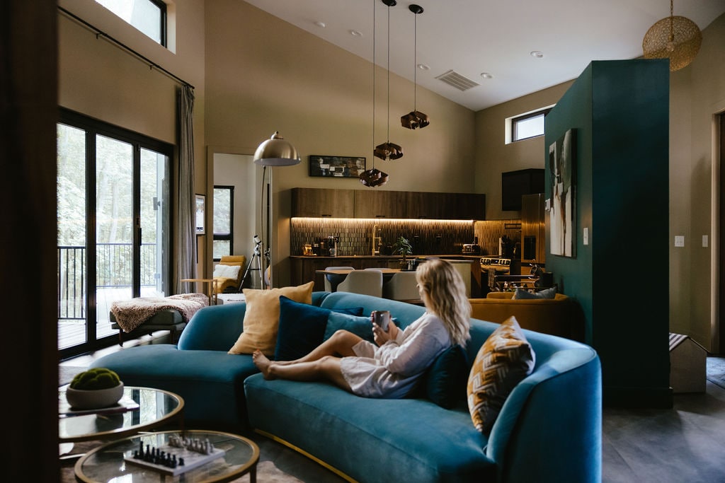 A spacious living area features soft blue seating complemented by vibrant cushions. Large glass doors provide access to a private balcony, while modern lighting illuminates the open kitchen area with wooden cabinetry. A person is seated comfortably, enjoying a drink and the serene ambiance.