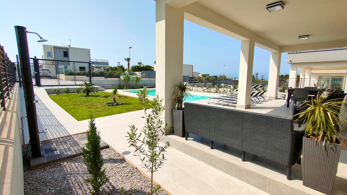 An outdoor area is showcased, featuring a modern patio furnished with comfortable seating. Lush greenery and potted plants frame the space, which leads to a sparkling pool. The property is enclosed, providing a sense of privacy, and offers ample sunlight.
